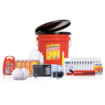 A survival emergency kit set arranged on a white background, featuring a red plastic bucket with a black toilet seat lid, food, water pouches, white masks, a black radio, a whistle, blanket, poncho, and first aid kit.