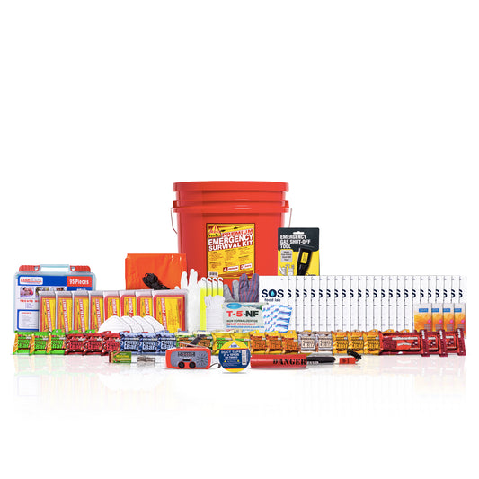 A comprehensive emergency survival kit arranged on a white reflective surface. The kit includes a large red bucket, an orange first-aid box, a blue and white battery, various yellow and orange food bars, water bottles, first-aid supplies, and other survival gear.