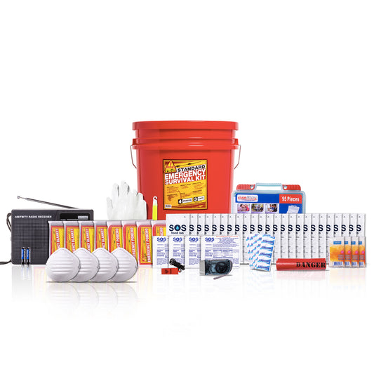 A comprehensive emergency survival kit arranged on a white reflective surface. The kit includes a large red bucket labeled 'STANDARD EMERGENCY SURVIVAL KIT', a black portable radio, three white respirator masks, several orange and yellow tubes, boxes of tablets, a small camera, a red fire starter, and other survival supplies.
