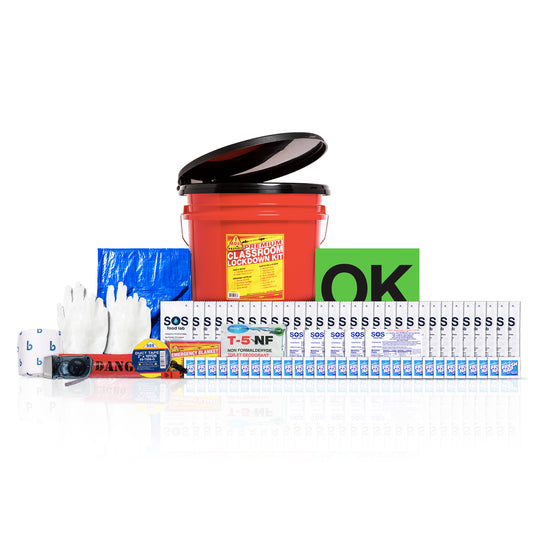 A classroom lockdown kit set displayed on a reflective white background, including a red bucket with a black lid, a green 'OK' sign, white safety gloves, blue gloves, a digital camera, a roll of tape, and multiple white lockout/tagout tape rolls.
